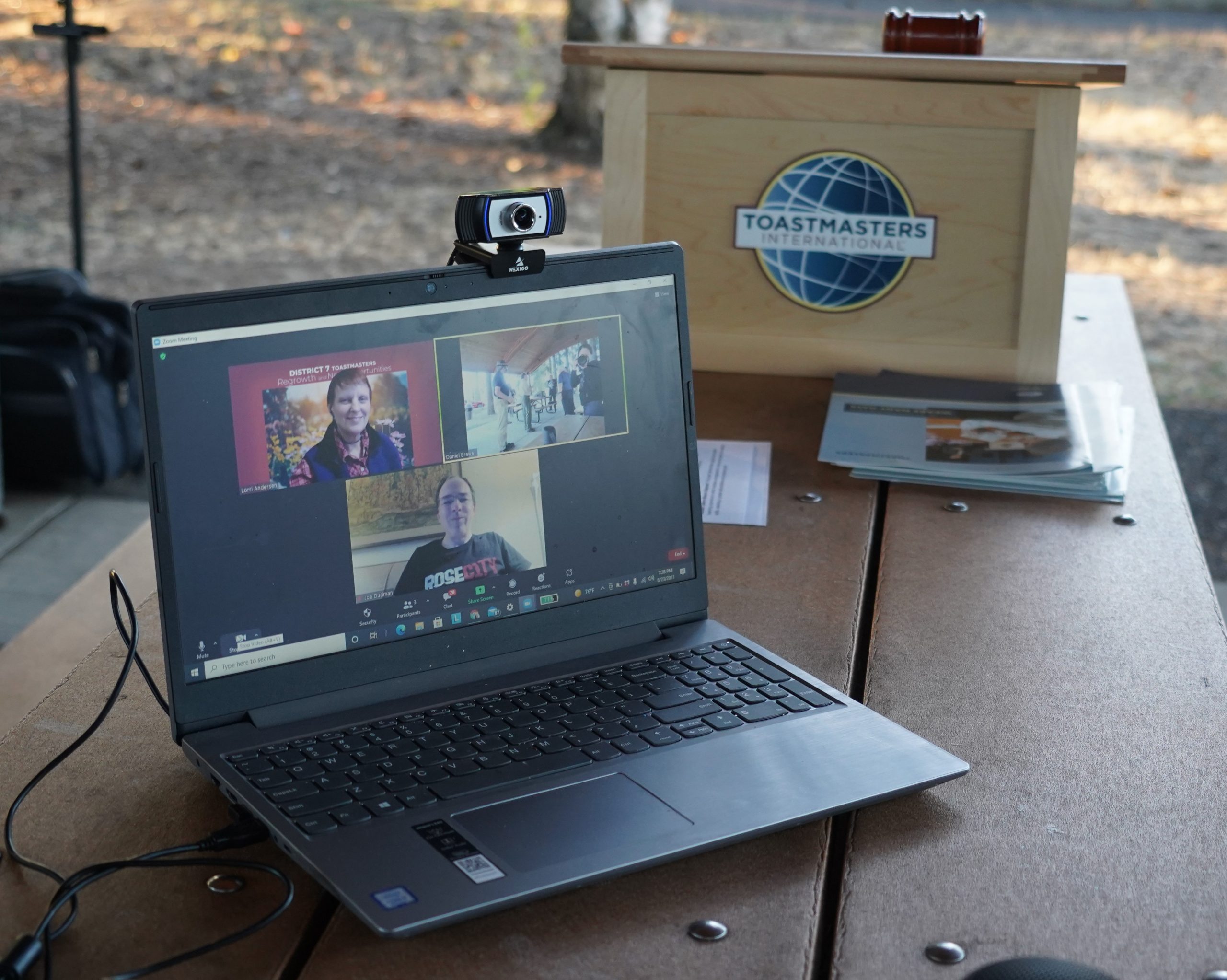 A laptop with web cam mounted, with several Zoom participants showing on screen, and Toastmasters portable lectern and gavel in the background